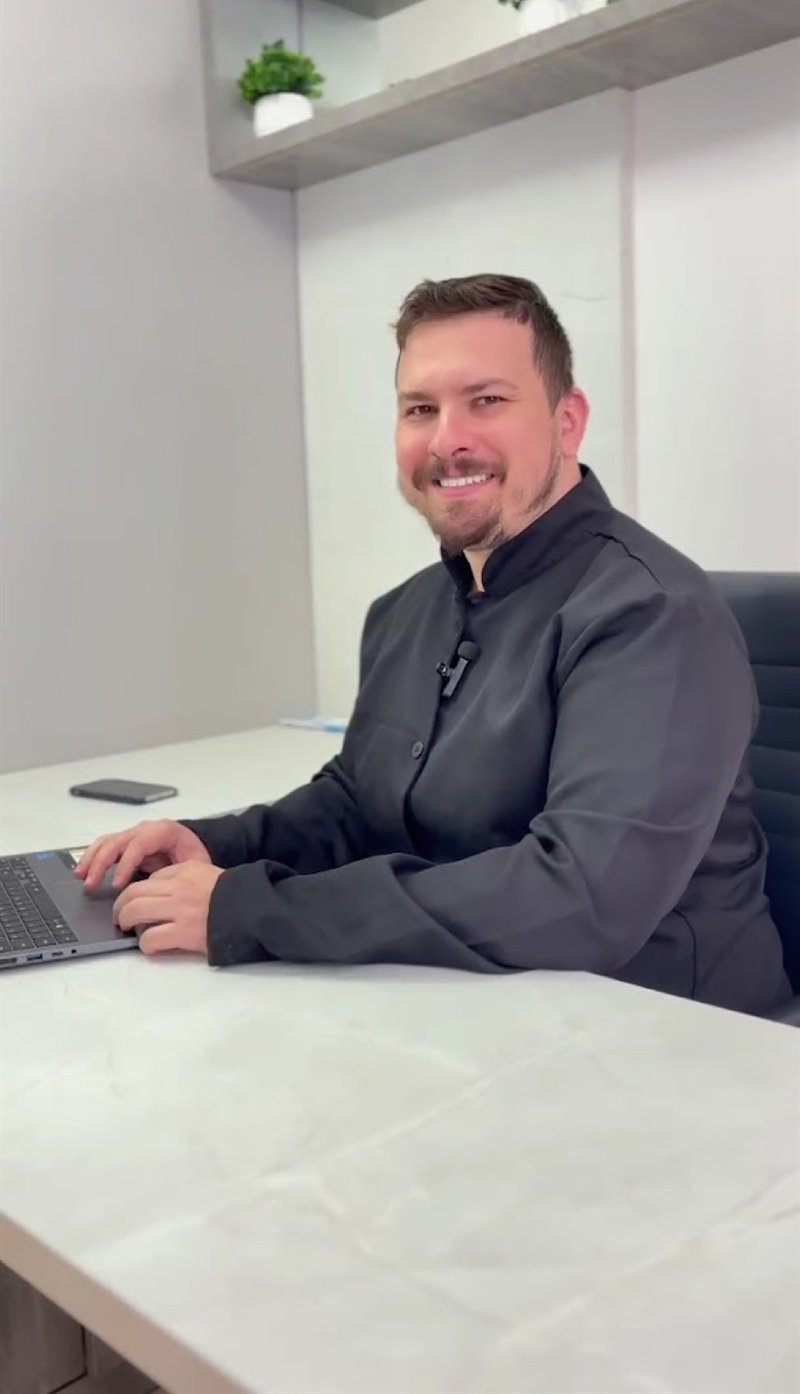 Dr. Kaio Cesar Grein atendendo em seu consultório, sorrindo à mesa de trabalho